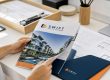 Moderner heller Schreibtisch mit buntem Hochglanz-Immobilienprospekt und gedruckten Grundrissplänen – professionelle Immobiliendokumentation