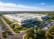 Vogelperspektive auf modernen Industriecampus mit Produktionshallen, Parkplatz, Zufahrtsstraße und grünen Freiflächen bei Sonnenlicht