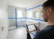 Mann mit Tablet vermisst modernes Badezimmer diagonal – blaue digitale Maßlinien zeigen präzise Abstände von Wänden, Dusche und Badewanne